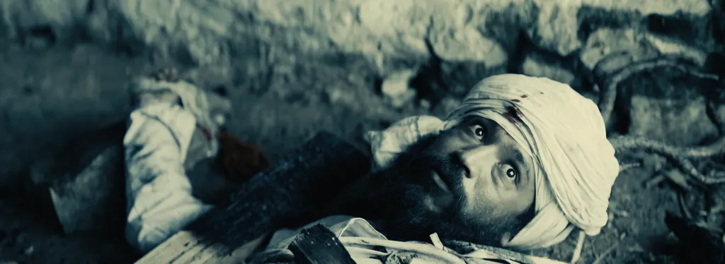 A wounded man in a white turban lying on rocky ground