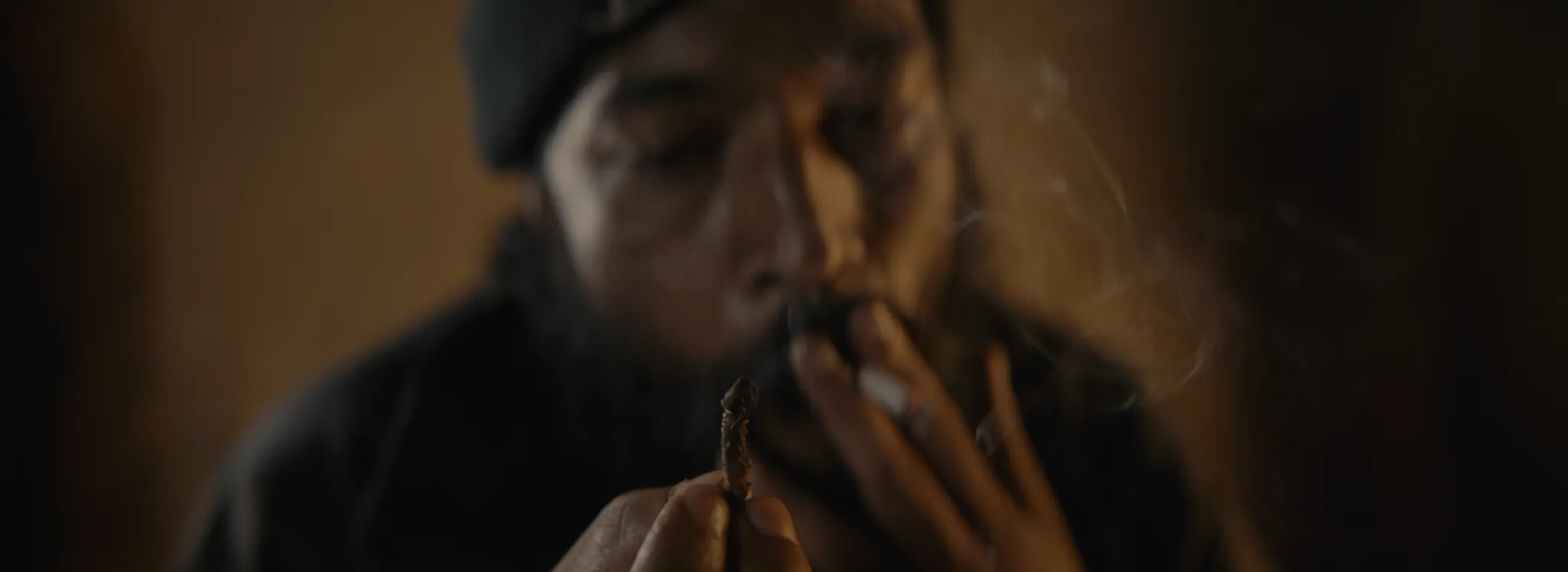 A man in a dark cap lighting a cigarette in warm light, from KRIMI