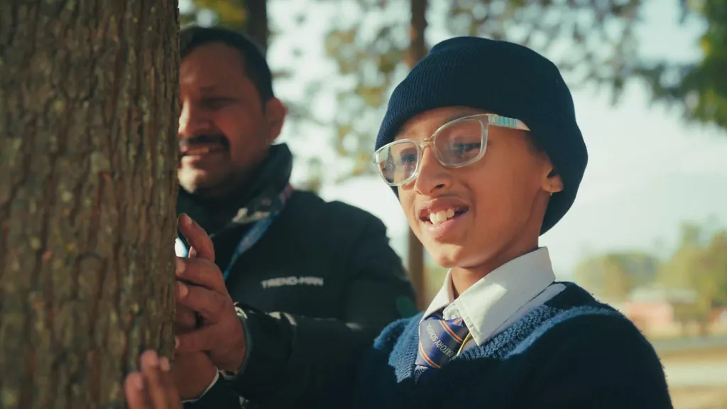 A schoolboy in a woollen hat touching a tree trunk while his teacher watches