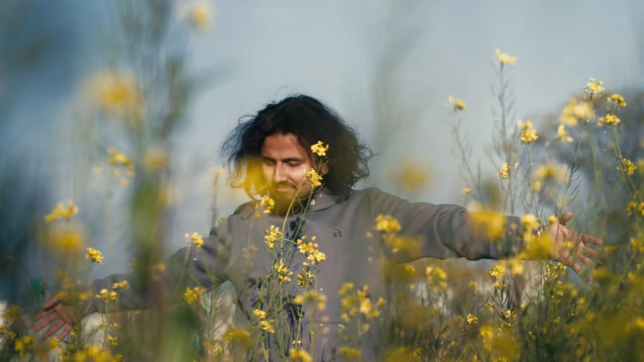 A man with long hair standing among yellow mustard flowers, from Madhur Jhankaar