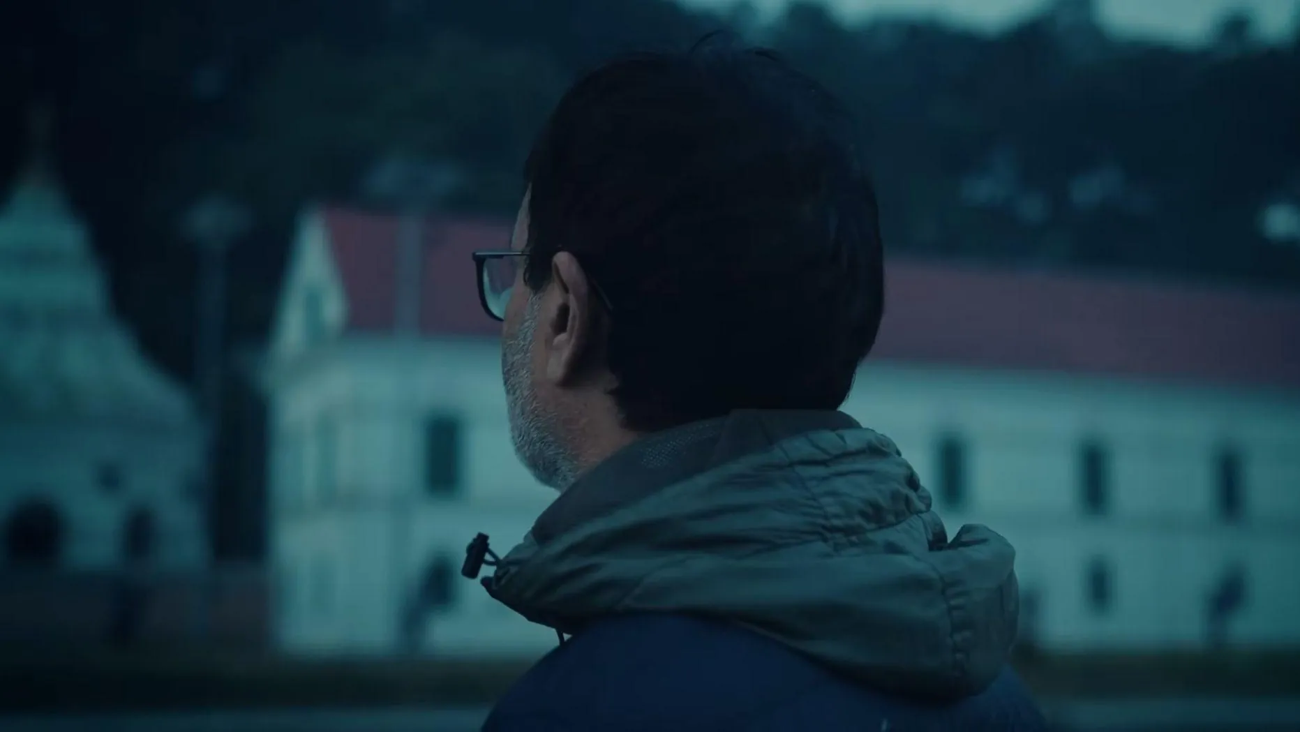 A man in a grey hood seen from behind, facing an old white building, from Samaya ko Cycle